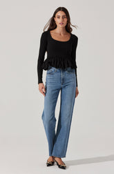 A woman is wearing a black long-sleeved top with a scoop neckline and a ruffled peplum hem. She is also wearing high-waisted, straight-leg blue jeans. Her shoes are black with a bow detail on the front and a small heel.